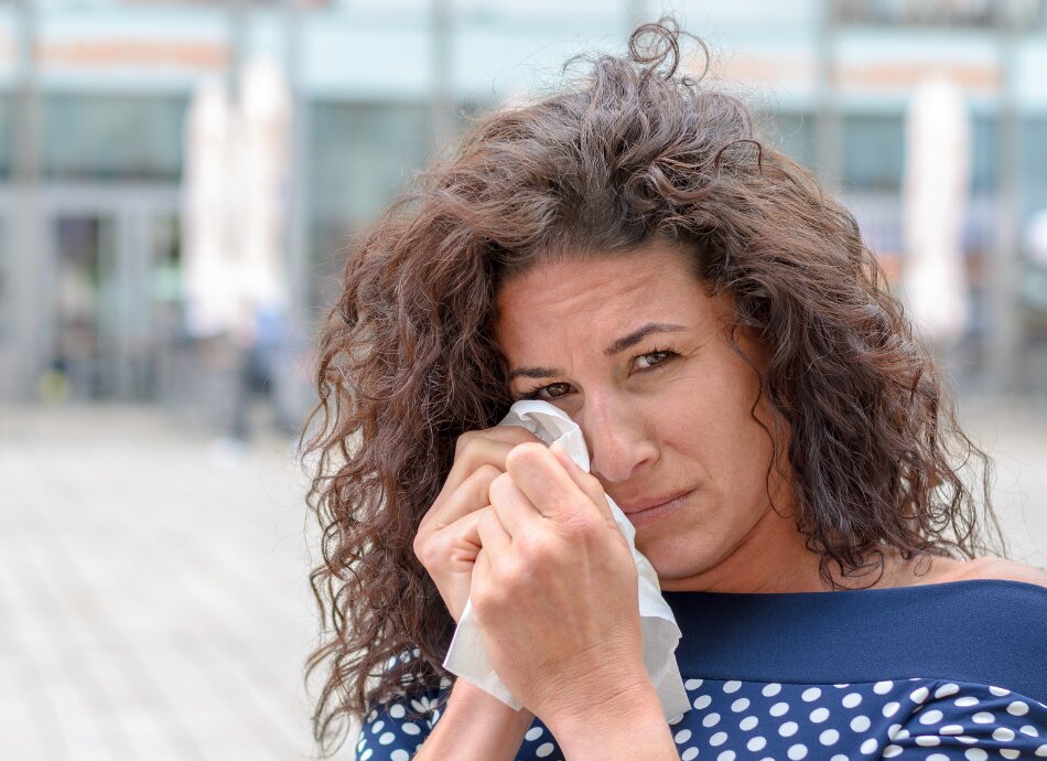 Curly-haired woman outdoors wiping eye with tissue