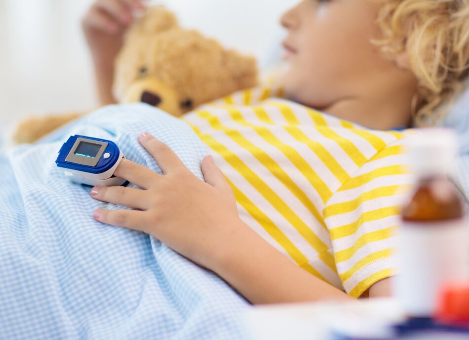 Child lies in bed holding teddy with pulse oximeter on left middle finger