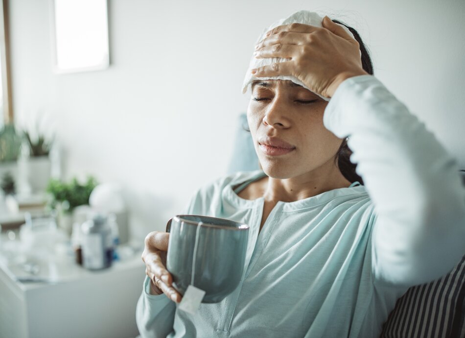 Unwell young woman holding teacup with towel on forehead