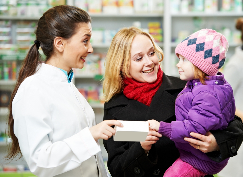 Pharmacist handing over white box of tablets to mother and toddler daughter in pharmacy Canva 950x690