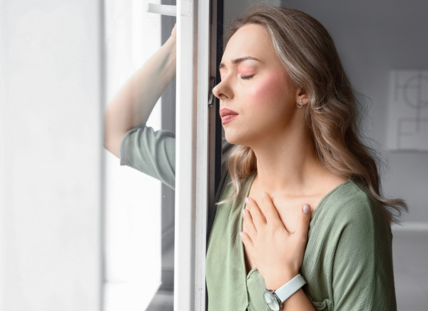 Young woman standing near window with hand on chest trying to stay calm