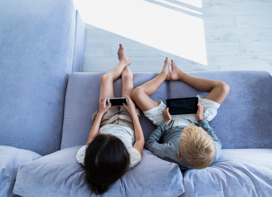 2 children lie on couch with cellphone and tablet