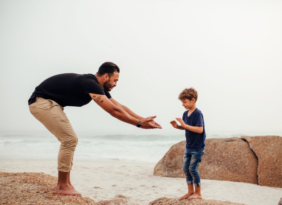 Man reaching hands out to boy to help jump from rock to rock on beach