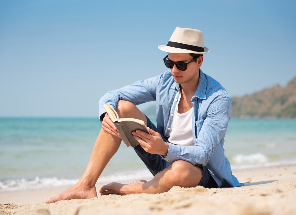 Man reading book on beach in hat and sunglasses
