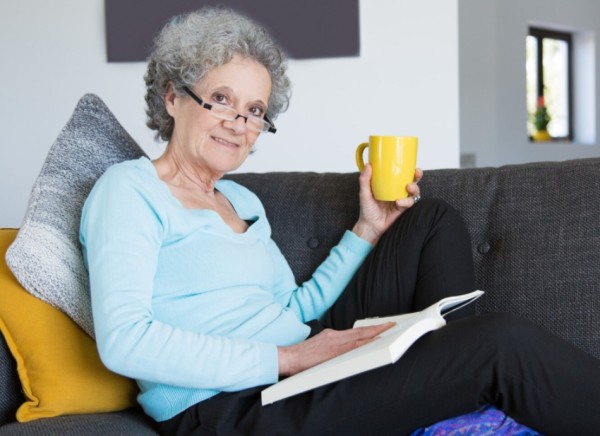 Slim older woman having hot drink while sitting on couch and reading